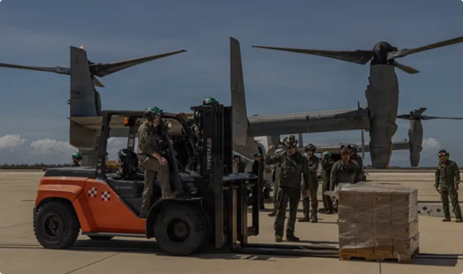 A forklift loading supply pallets beside a tiltrotor aircraft.
