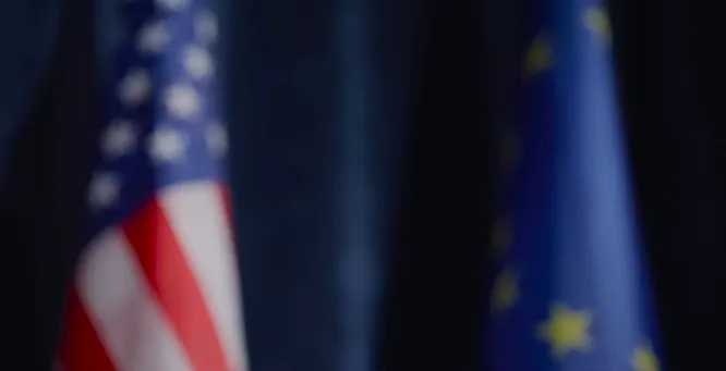 Two officials shaking hands in front of American and European Union flags.